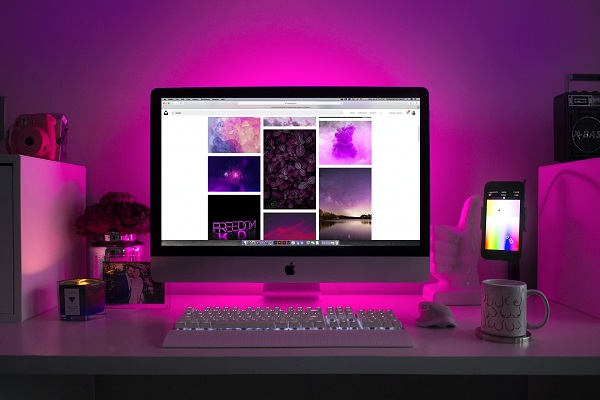 A desk with a desktop computer, keyboard, and a monitor displaying images, with a mug, mobile phone, flowers, and a photo